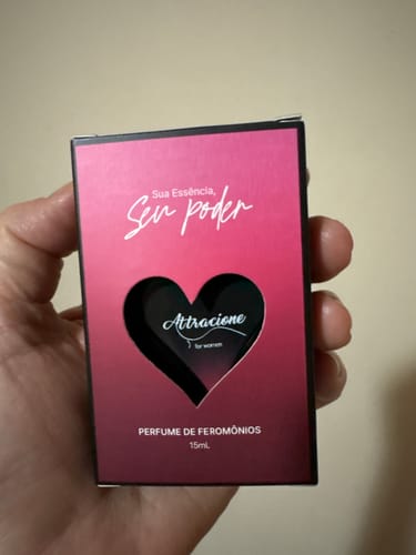 Reviewer's hand holding a small pink and dark red box of Attracione Sensual perfume with a heart-shaped cutout.