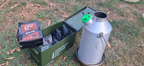 Customer's Coffee Resupply with four coffee bags in an open green metal ammo tin next to a silver metal kettle outside.