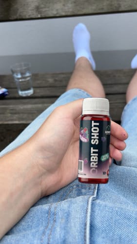 Reviewer's hand holding a red Orbit Shot bottle while relaxing on a wooden bench.