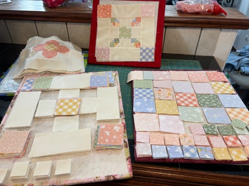 Reviewer's progress on a Garden Gate Quilt Kit with various pastel fabric squares and blocks laid out on work surfaces.