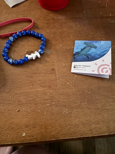 Customer's Shark Week Bracelet with blue beads and hammerhead charm, shown with an informational card on a wooden table.