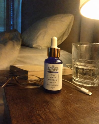 Customer's Advanced Peptide serum bottle on a bedside table with a book, glasses, and a glass of water.