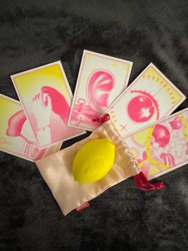 Customer's yellow Lem clitoral massager sitting in a satin drawstring pouch surrounded by several colorful art cards.