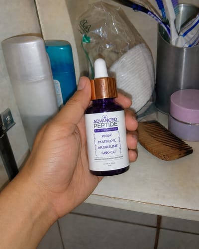 Reviewer's hand holding the Advanced Peptide serum bottle with a white dropper cap in a bathroom setting.