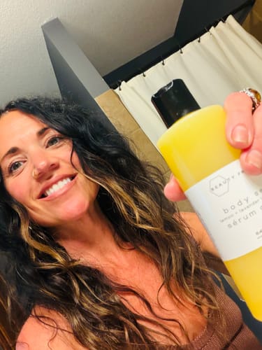 Customer smiling while holding a yellow bottle of Body Serum in a bathroom setting.