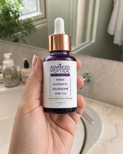 Reviewer's hand holding a purple Advanced Peptide serum bottle with a white dropper lid in a bathroom.