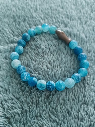 Reviewer's blue, crackled bead Voyage Bracelet with a small shark charm, placed on a textured blue fabric surface.