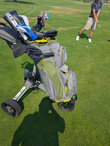 Customer's Cybercart golf caddy with a grey bag on a golf course while a person prepares to hit a ball nearby.