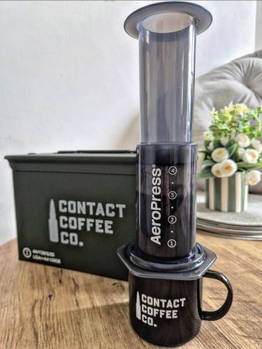 Customer's black enamel mug with Contact Coffee logo, shown with an AeroPress coffee maker on a table.