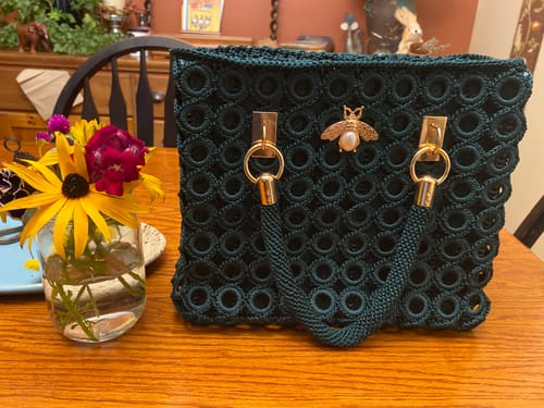 Customer's completed dark green Garden Bee Crochet Bag with a golden bee charm sitting on a wooden table near flowers.