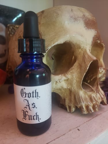 Customer's blue dropper bottle of July 5: Your Choice Hair and Body Oil with Goth As Fuck label, beside a skull.