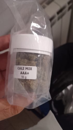 Customer's hand holding a sealed plastic bag containing a small jar of CALI MIX AAA+ flowers.