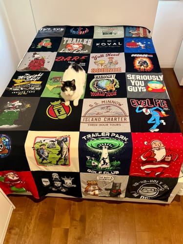 Customer's Project Repat T-Shirt Quilt featuring various graphic squares, with a cat sitting in the center on a bed.