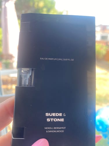 Customer's hand holding a black 2ml Suede & Stone Eau de Parfum sample bottle outdoors.