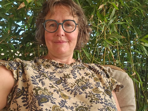 Reviewer porte la blouse Camélia avec son imprimé floral, son col rond volanté et ses manches kimono devant des bambous.