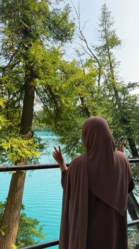 Reviewer wearing a brown Klaythelabel hijab, posing with peace signs in front of a lake and trees.