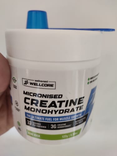 Customer's hand holding a white Wellcore Creatine Monohydrate container with a blue scoop on top.