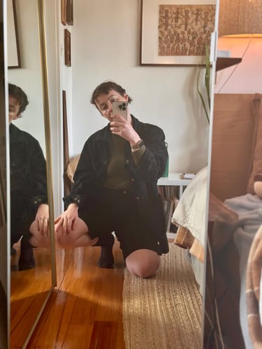 Customer's Long Shorts in a dark color, worn while crouching and taking a mirror selfie.