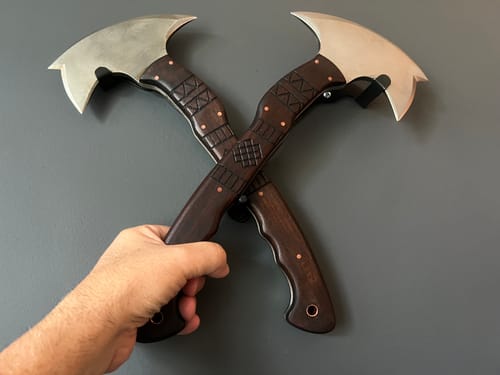 Customer's hand holding two crossed tomahawks displayed on the metal wall mount.
