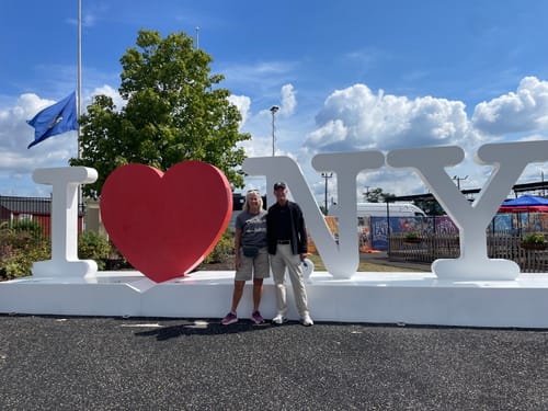 Customer wearing a Clowns and Jokers Basic T-Shirt while standing in front of an I Love New York sign at a fair.