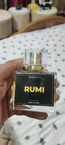 Customer's hand holding a clear, rectangular bottle of Rumi perfume with a gold cap and a black label.
