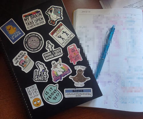 Reviewer's black Kristy Planner covered in quirky stickers next to a blue pen on an open calendar page