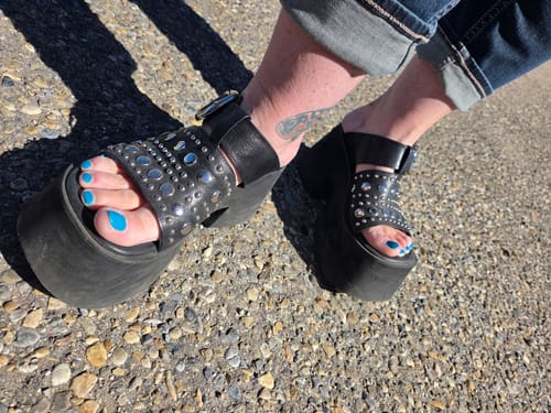 Customer's black leather studded platform slippers being worn on a gravel surface.