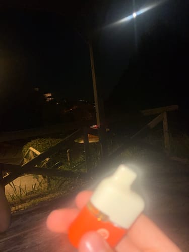 Customer's hand holding an orange THC-A Vape at night, in a blurry, dimly lit outdoor setting.