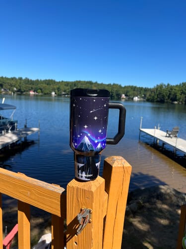 Customer's Thicc Buddy mug with a night sky design perched on a wooden fence overlooking a scenic lake.