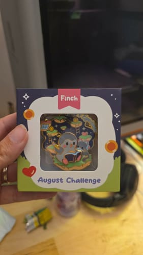 Customer's August Challenge Finch enamel pin in original product packaging box held in a hand.