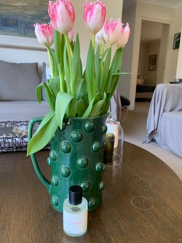 Customer's Lucid Dreaming fragrance bottle placed on a wooden table beside a green textured vase with pink tulips.