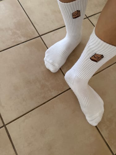 Customer's white UGLYFACE® CLASSIC SOCKS with embroidered brown bag logo, worn on feet over tiled floor.