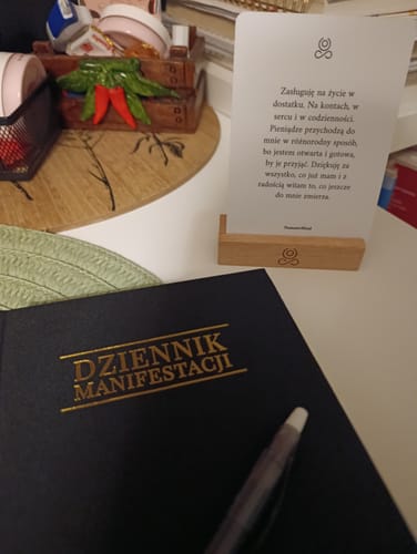 Reviewer's czarny Dziennik Manifestacji z długopisem na biurku obok karty z afirmacją w drewnianym stojaku