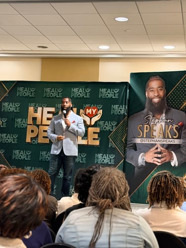 Customer's view of a speaker on stage at the Heal My People Tour, with event branding in the background.