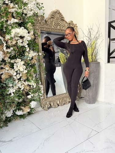 Reviewer standing in a black bodycon Naked Off-Shoulder Closed Toe Jumpsuit in front of a mirror with floral decor.