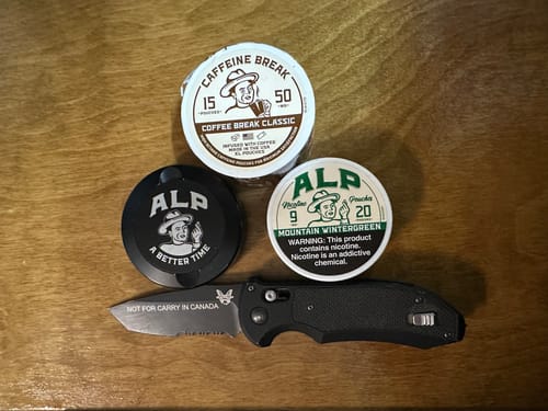 Customer's Coffee Break Caffeine Pouches tin alongside two other round tins and a black folding knife on a wooden surface.