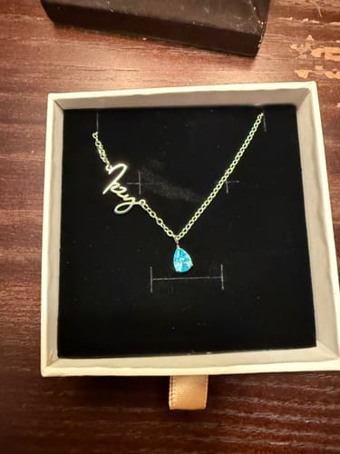 Customer's Lucce Signature Necklace with a script name and a blue teardrop stone, displayed inside its gift box.