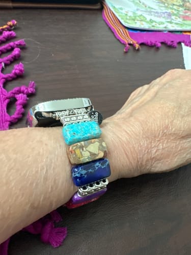 Customer's hand wearing a Chic Chakra Stretch Apple Watch Strap with colorful stone segments on a dark table surface.