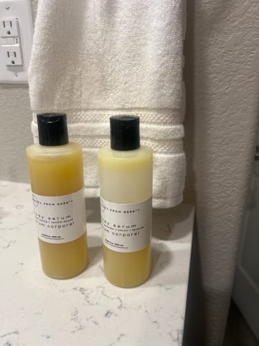 Customer's two Body Serum bottles, one yellow and one light yellow, standing on a bathroom counter near a towel.