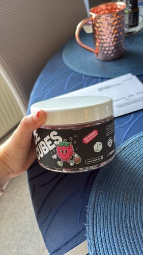 Reviewer's Hand hält eine Dose THC Space Cubes mit Himbeer-Illustration auf einem blauen Tisch.