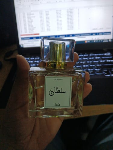 Customer's hand holding a square bottle of Sultan perfume with a gold cap in front of a laptop screen.