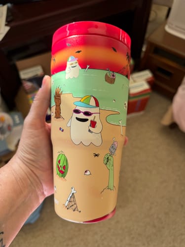 Reviewer holding the To-Go Buddy cup featuring a colorful beach-themed ghost print against a sunset background.