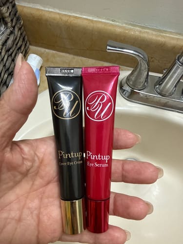 Reviewer holding a black Meishoku Pintup Cover Eye Cream tube next to a red eye serum tube in a bathroom setting.