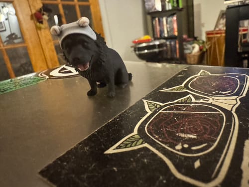 Customer's DoDoWish Custom 3D Pet Figurine, a black dog wearing a small grey hat, sitting on a table.