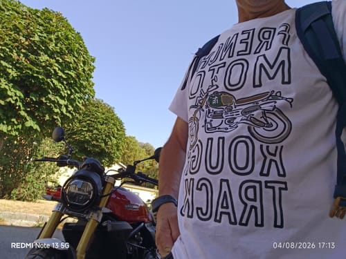 Client portant le t-shirt blanc Collection Mc Queen à côté d'une moto rouge dans un cadre arboré.