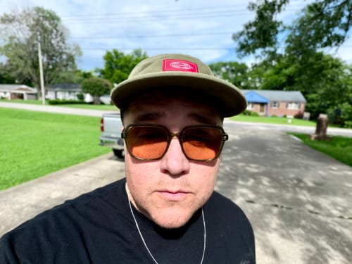 Reviewer wearing a green hat with a red logo patch and tinted sunglasses, outdoors.