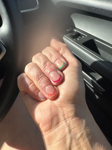 Reviewer's hand wearing Holy Gels with a thin, colorful French tip design on each nail while sitting in a car