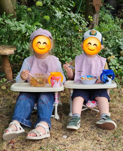 Customer's children eating while seated in two Paloma baby cotton hat high chairs outdoors in a garden setting