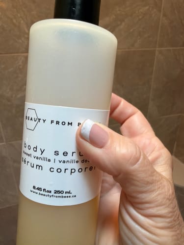 Customer's hand holding a bottle of Sweet Vanilla Body Serum in a tiled shower.