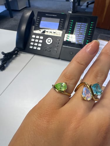 Customer's hand wearing two gold-toned Pia Birthstone Rings, one with a pear-cut green stone and one with blue stones.